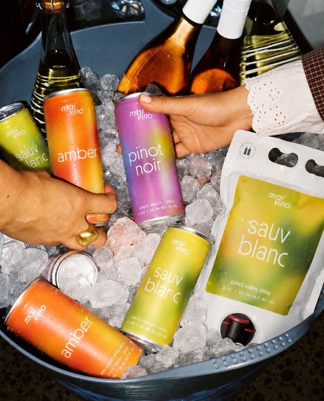 Hands reaching for Mai Vino Amber, Pinot Noir and Sauv Blanc cans and a Sauv Blanc pouch in a bucket of ice